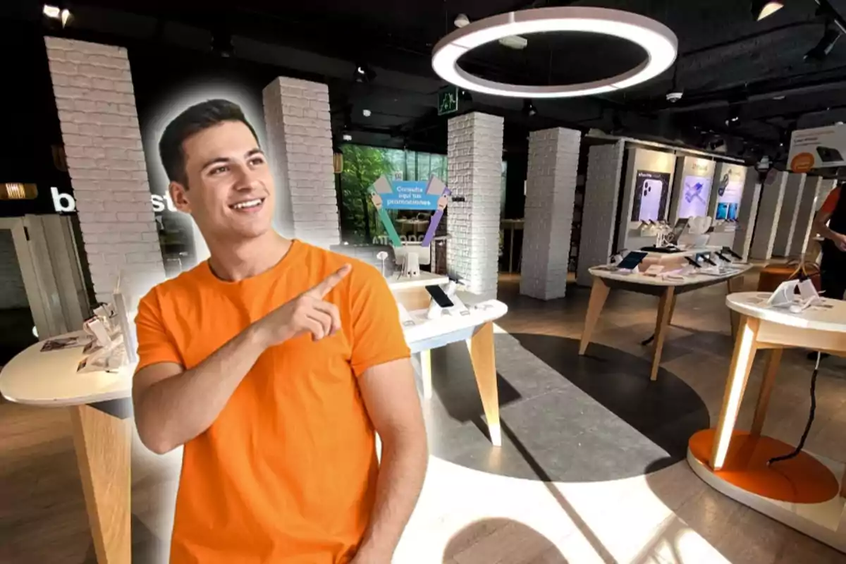 Un hombre con camiseta naranja está sonriendo y señalando con el dedo en una tienda de tecnología moderna con mesas que exhiben dispositivos electrónicos.