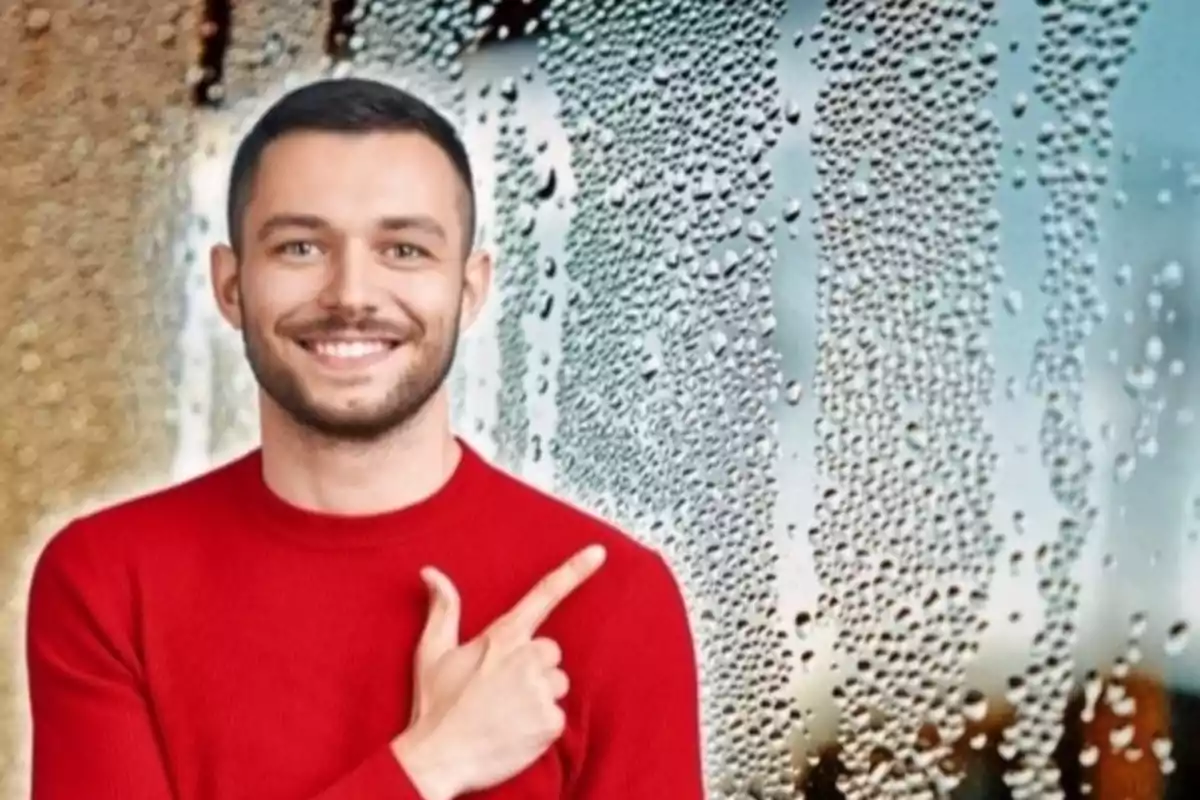 Hombre con suéter rojo sonriendo y señalando hacia la derecha, con un fondo de ventana con gotas de agua.