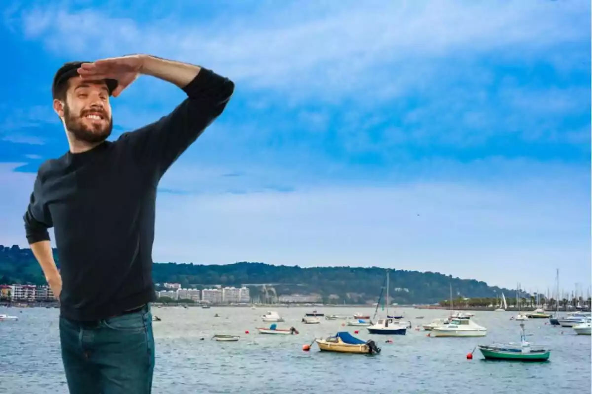 Un hombre sonriente con barba y camiseta negra está de pie junto a un puerto, mirando al horizonte con la mano en la frente como si estuviera buscando algo, mientras varios barcos están anclados en el agua y un cielo azul con algunas nubes se extiende sobre el paisaje.