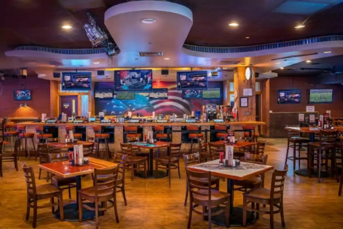 Sports bar-style restaurant with wooden tables and chairs, several televisions turned on, and a bar in the back decorated with a mural of the American flag. Sports bar-style restaurant with wooden tables and chairs, several televisions turned on, and a bar in the back decorated with a mural of the American flag.