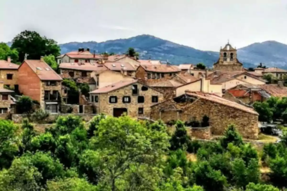 Vista panorámica de un pequeño pueblo con casas de piedra y techos de tejas rojas rodeado de vegetación y montañas al fondo.