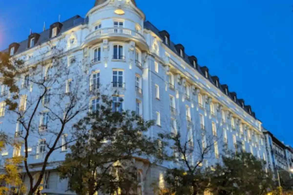 Edificio blanco de varios pisos con ventanas iluminadas y árboles en primer plano durante el atardecer.