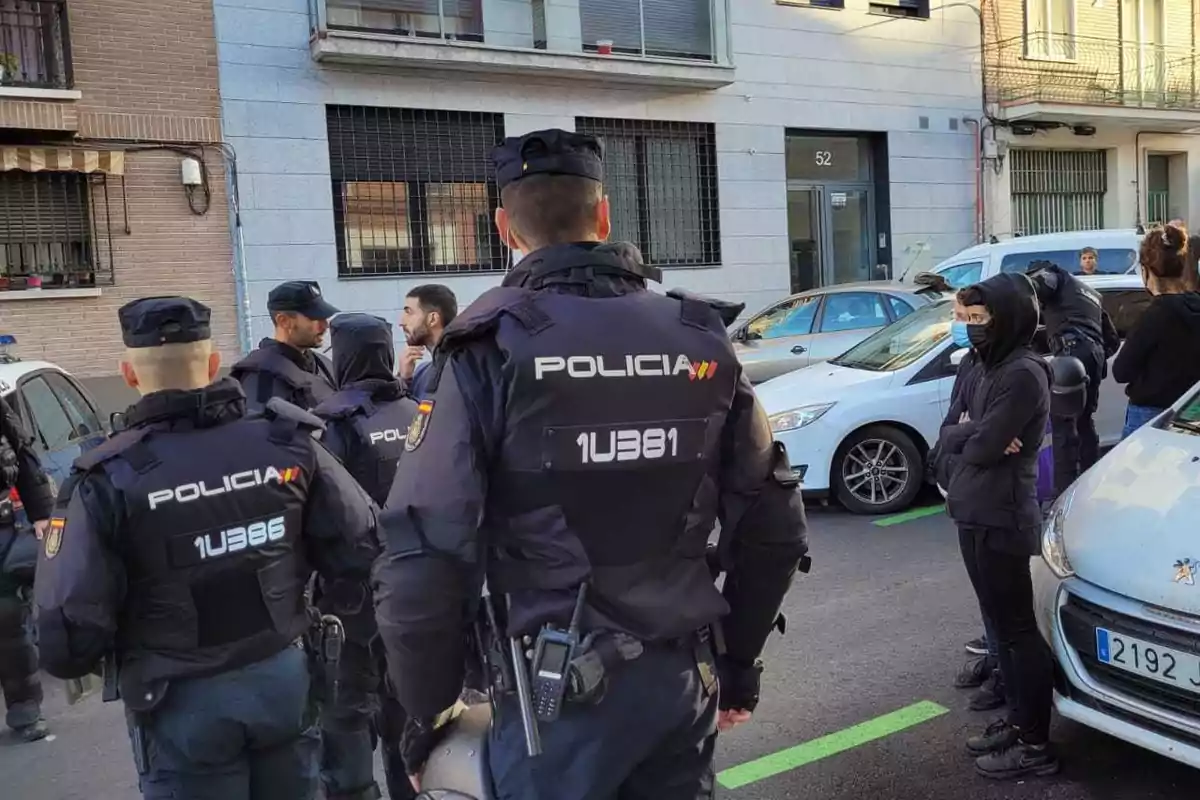 Un grupo de policías con uniformes oscuros está en una calle junto a varios vehículos y personas, algunos de los cuales llevan mascarillas.