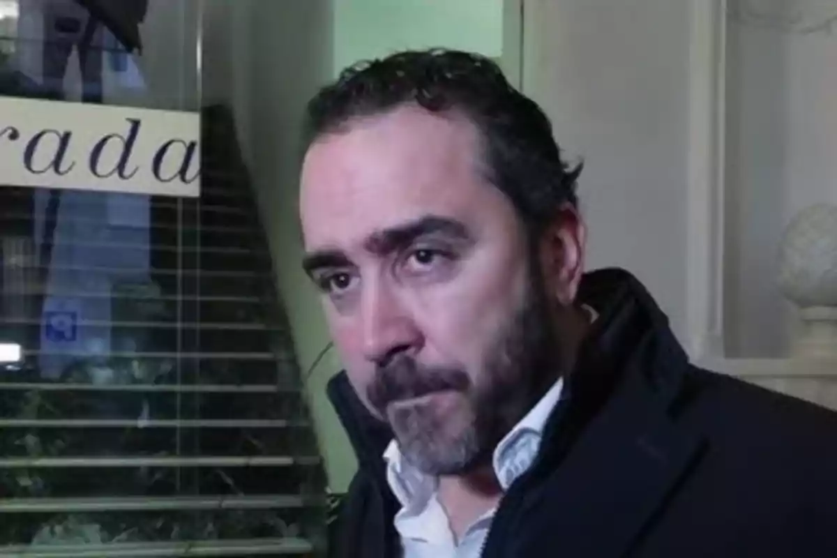 A man with a beard and dark hair is standing in front of a staircase in an indoor setting.