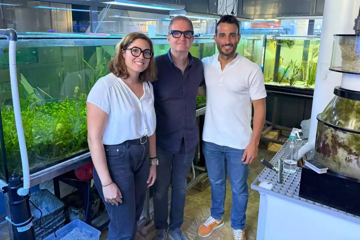 Tres personas posan sonrientes en un laboratorio de acuarios rodeados de tanques con plantas acuáticas y equipo científico