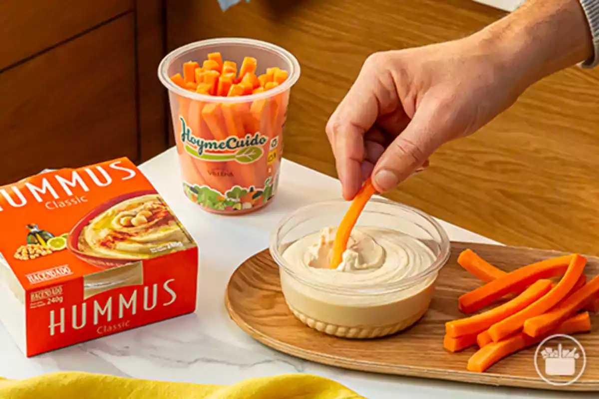 Una mano sumerge un palito de zanahoria en un recipiente de hummus junto a una bandeja con más zanahorias y envases de hummus y zanahorias en la mesa Una mano sumerge un palito de zanahoria en un recipiente de hummus junto a una bandeja con más zanahorias y envases de hummus y zanahorias en la mesa