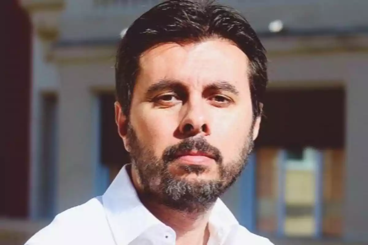 Un hombre con barba y cabello oscuro lleva una camisa blanca y está al aire libre con un edificio desenfocado de fondo.