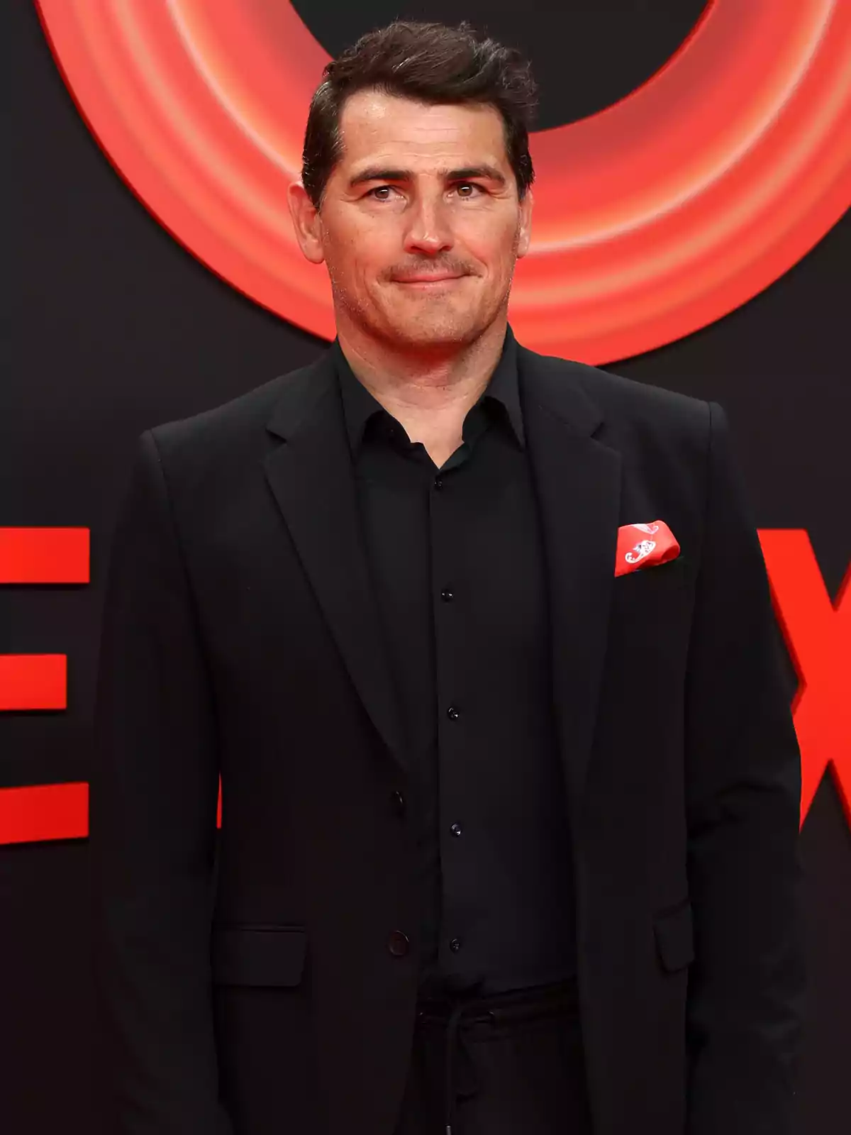 Iker Casillas vistiendo un traje negro con camisa oscura y pañuelo rojo en la solapa, posando frente a un fondo negro con detalles rojos. Iker Casillas vistiendo un traje negro con camisa oscura y pañuelo rojo en la solapa, posando frente a un fondo negro con detalles rojos.