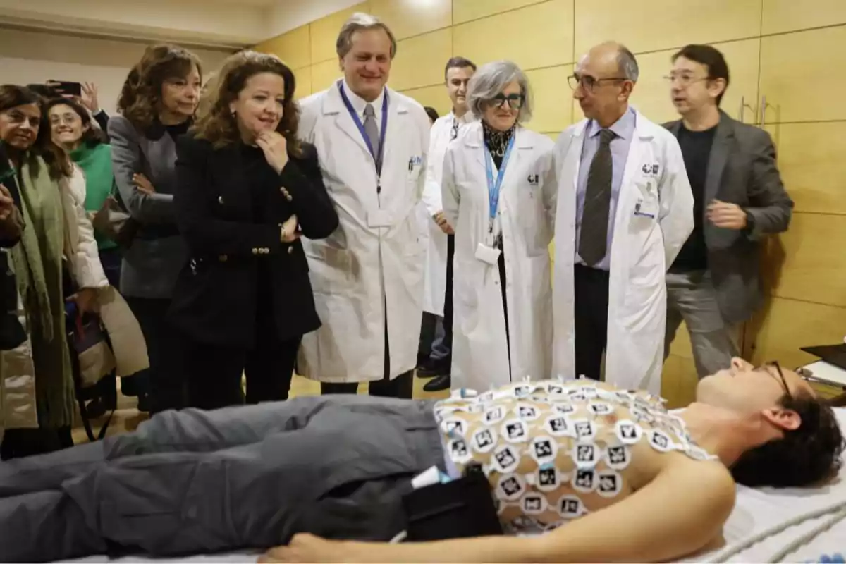 A group of people, some in white coats, watch a man lying on a stretcher with sensors on his chest.