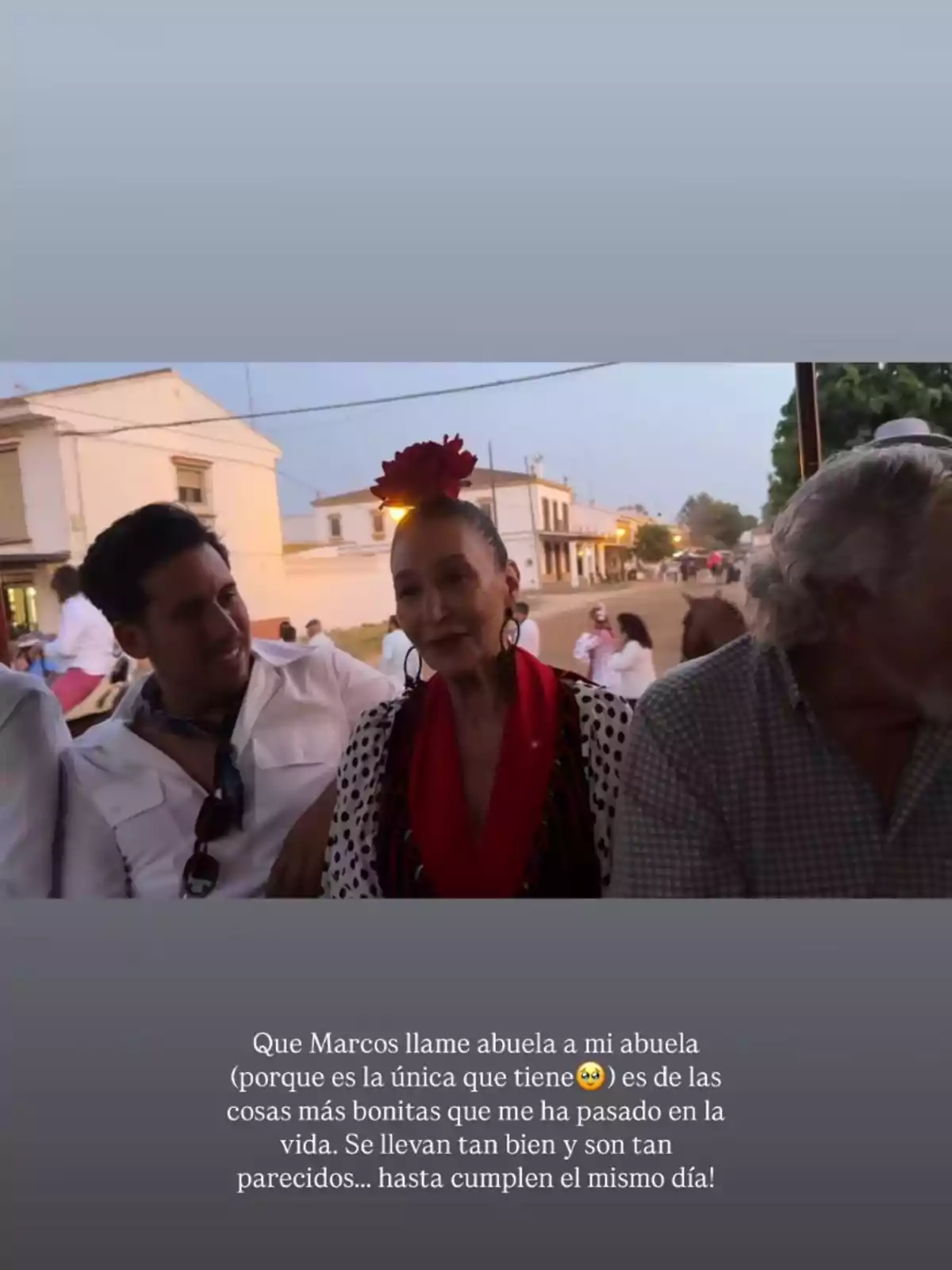 Tres personas sentadas juntas al aire libre, una mujer en el centro lleva una flor roja en la cabeza y viste ropa tradicional, mientras los hombres a su lado la acompañan sonrientes, al fondo se ven casas y más personas en la calle. Tres personas sentadas juntas al aire libre, una mujer en el centro lleva una flor roja en la cabeza y viste ropa tradicional, mientras los hombres a su lado la acompañan sonrientes, al fondo se ven casas y más personas en la calle.