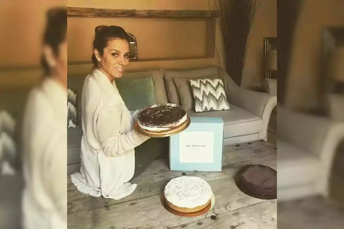 Una persona sentada en el suelo de una sala sostiene un pastel mientras sonríe a la cámara, con una caja azul y otros pasteles sobre una mesa de madera frente a un sofá. Una persona sentada en el suelo de una sala sostiene un pastel mientras sonríe a la cámara, con una caja azul y otros pasteles sobre una mesa de madera frente a un sofá.