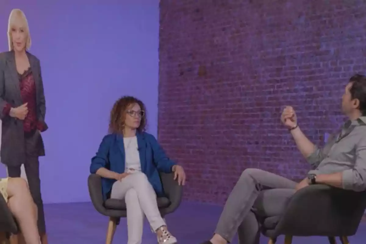 Un grupo de personas sentadas en sillas modernas conversan en una habitación con una pared de ladrillo y un fondo iluminado en tonos morados, mientras una figura de cartón de una mujer rubia está de pie a un lado. Un grupo de personas sentadas en sillas modernas conversan en una habitación con una pared de ladrillo y un fondo iluminado en tonos morados, mientras una figura de cartón de una mujer rubia está de pie a un lado.