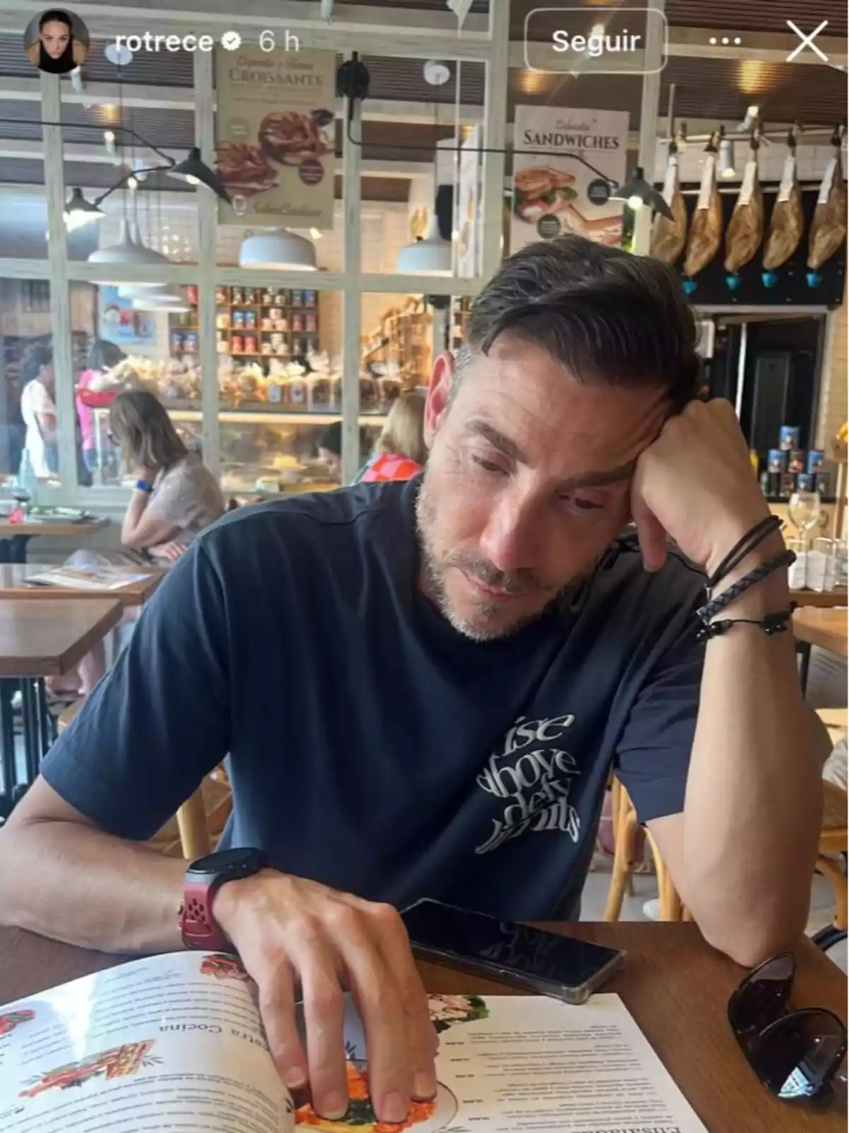 Antonio David Flores sentado en una cafetería mirando el menú con expresión pensativa, lleva una camiseta oscura y tiene un reloj rojo en la muñeca, sobre la mesa hay un par de gafas de sol y un teléfono móvil. Antonio David Flores sentado en una cafetería mirando el menú con expresión pensativa, lleva una camiseta oscura y tiene un reloj rojo en la muñeca, sobre la mesa hay un par de gafas de sol y un teléfono móvil.