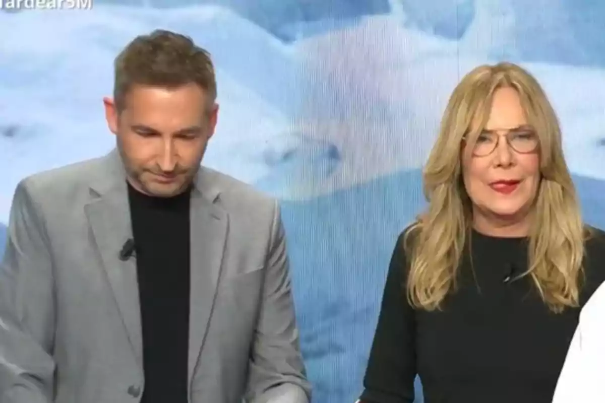 Frank Blanco y Belén Rodríguez están de pie frente a una pantalla azul, Frank lleva un traje gris y Belén un vestido negro. Frank Blanco y Belén Rodríguez están de pie frente a una pantalla azul, Frank lleva un traje gris y Belén un vestido negro.