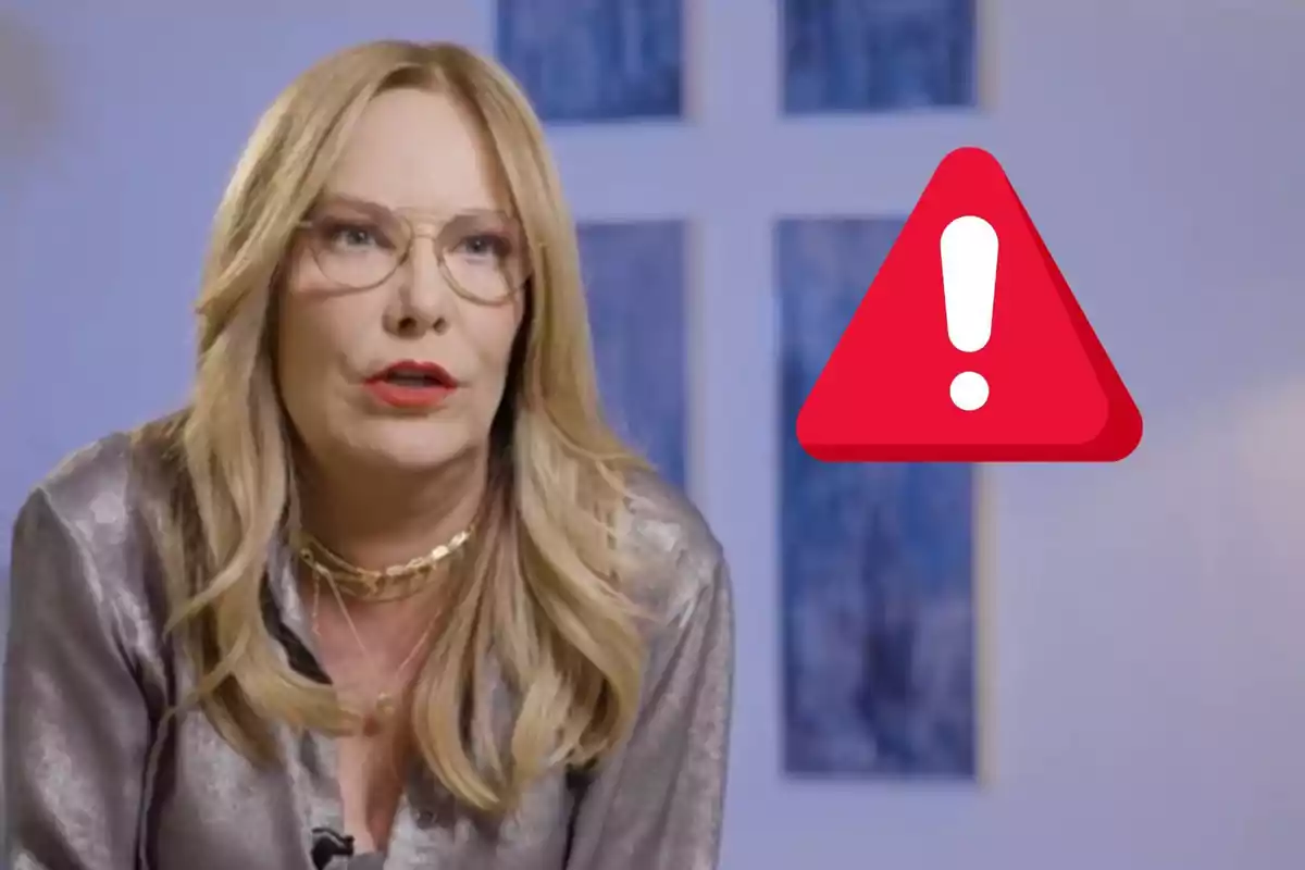 Belén Rodríguez con cabello rubio y gafas está hablando frente a un fondo azul con un símbolo de advertencia rojo al lado.