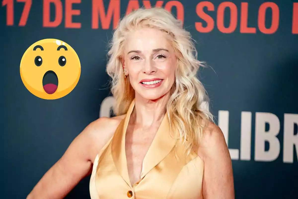 Belén Rueda rubia sonríe frente a un fondo con texto rojo y negro, acompañada de un emoji sorprendido.