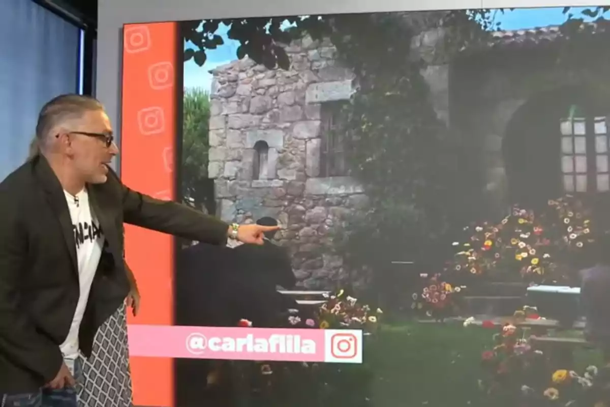 Kiko Hernández con gafas y chaqueta oscura señalando una pantalla grande que muestra una casa de piedra rodeada de flores y vegetación Kiko Hernández con gafas y chaqueta oscura señalando una pantalla grande que muestra una casa de piedra rodeada de flores y vegetación
