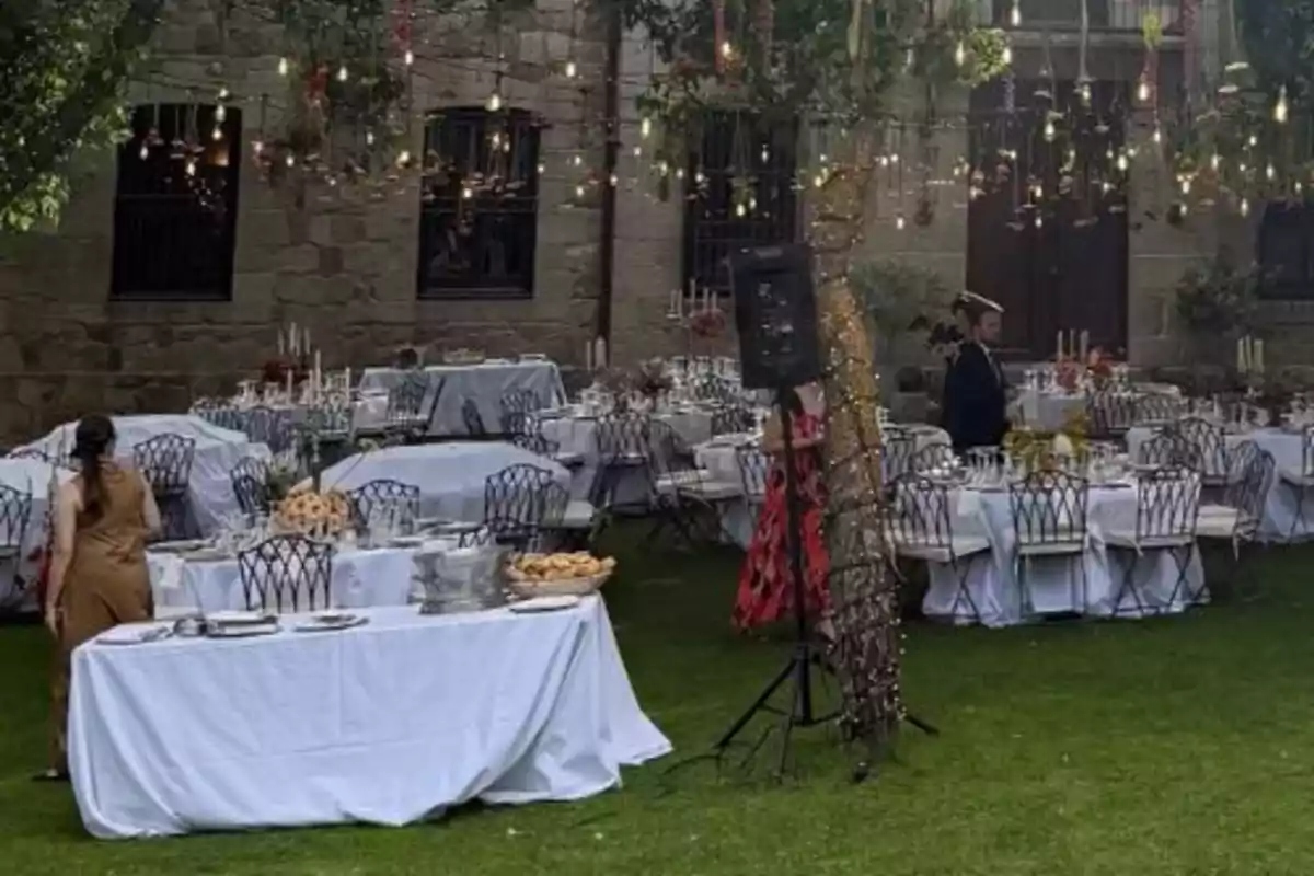 Evento al aire libre con mesas decoradas, manteles blancos, sillas metálicas y luces colgantes en un jardín junto a una pared de piedra Evento al aire libre con mesas decoradas, manteles blancos, sillas metálicas y luces colgantes en un jardín junto a una pared de piedra
