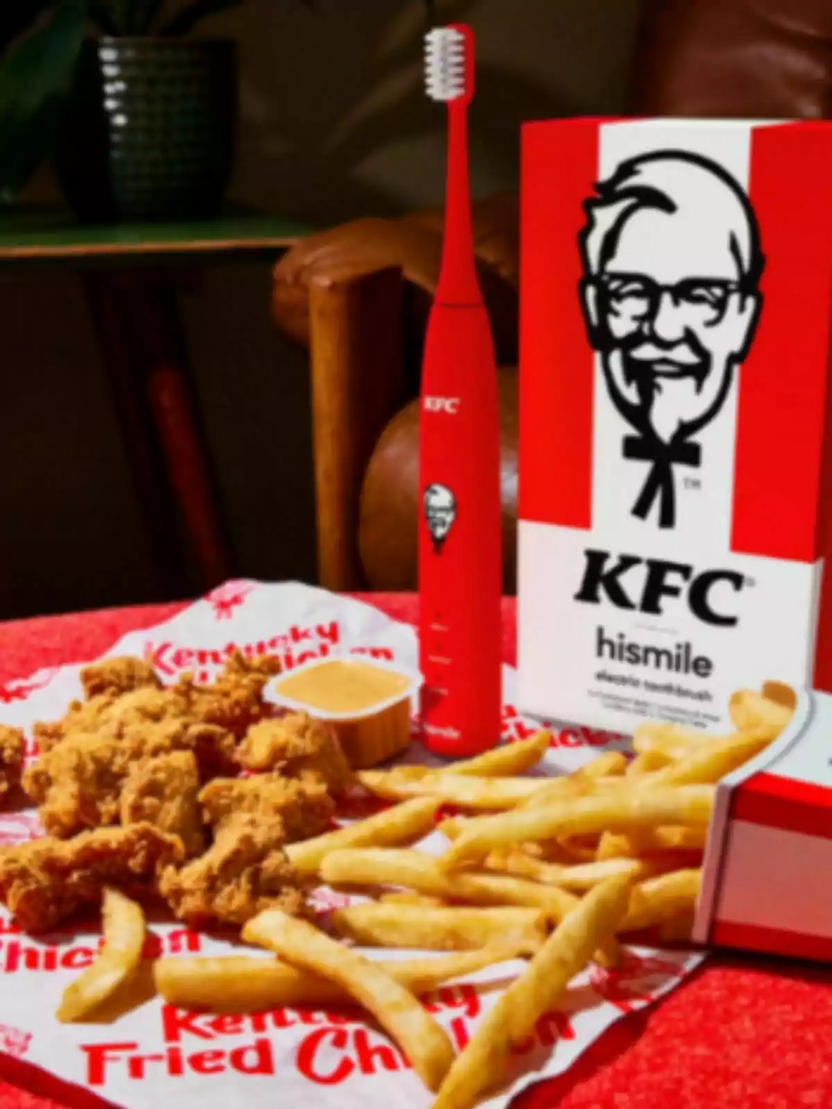 A red KFC electric toothbrush next to a box with the brand's logo, accompanied by a portion of fried chicken and French fries on a napkin with the name Kentucky Fried Chicken. A red KFC electric toothbrush next to a box with the brand's logo, accompanied by a portion of fried chicken and French fries on a napkin with the name Kentucky Fried Chicken.