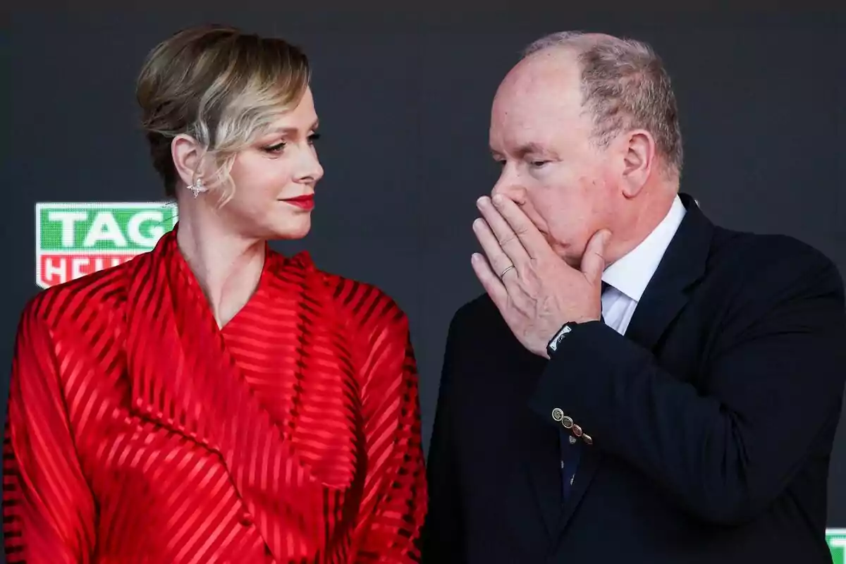A woman in a red dress looks at a man in a dark suit who covers his mouth with his hand while speaking to her at a formal event.