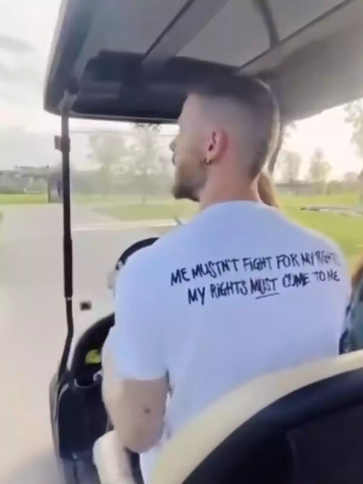 David de Gea conduce un carrito de golf en un entorno al aire libre, lleva una camiseta blanca con un mensaje en la espalda. David de Gea conduce un carrito de golf en un entorno al aire libre, lleva una camiseta blanca con un mensaje en la espalda.