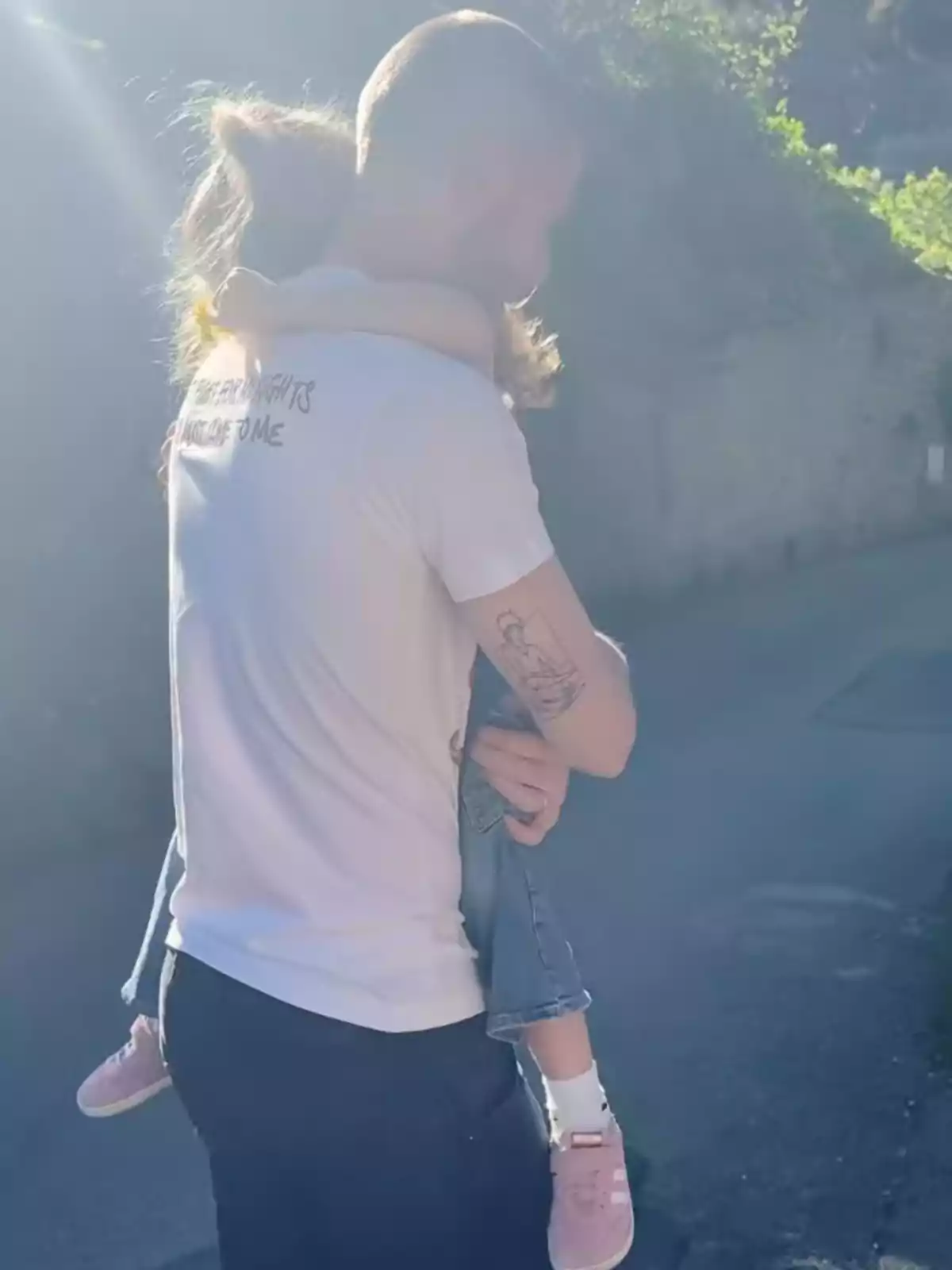 David de Gea con camiseta blanca y tatuaje en el brazo sostiene a una niña con zapatillas rosas en un camino iluminado por el sol. David de Gea con camiseta blanca y tatuaje en el brazo sostiene a una niña con zapatillas rosas en un camino iluminado por el sol.