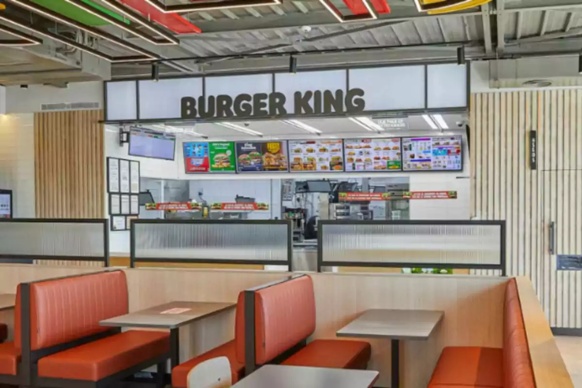 Interior de un restaurante de Burger King con mesas y sillas de color rojo y gris, un mostrador al fondo con menús iluminados y un letrero grande en la parte superior. Interior de un restaurante de Burger King con mesas y sillas de color rojo y gris, un mostrador al fondo con menús iluminados y un letrero grande en la parte superior.