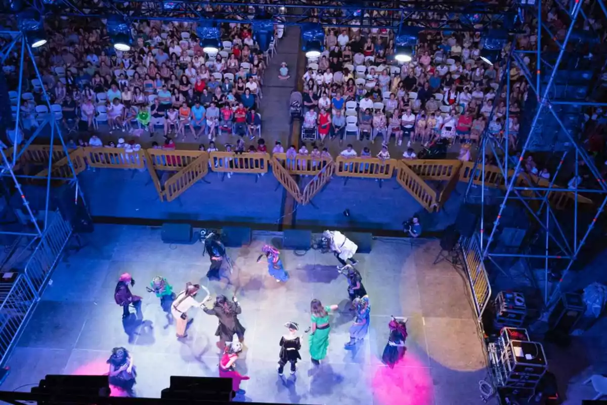 Un grupo de personas disfrazadas realiza una actuación en un escenario iluminado, mientras una gran audiencia observa desde sus asientos.