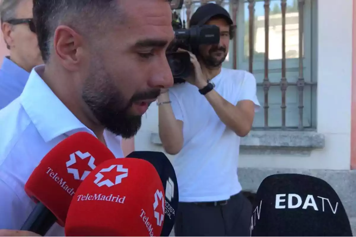 Un hombre con barba y camisa blanca está siendo entrevistado por varios medios de comunicación, mientras un camarógrafo lo graba.