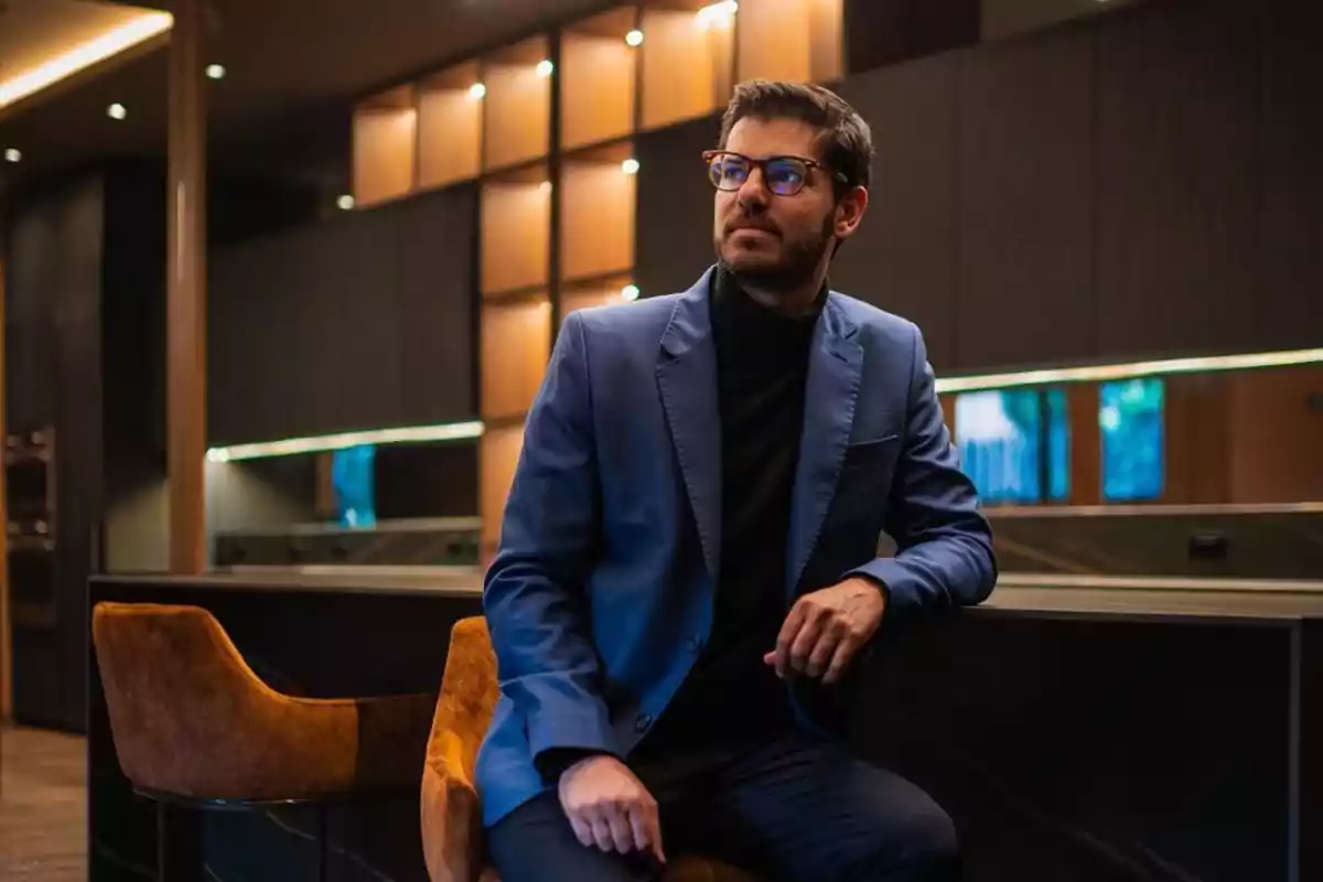 Un hombre con gafas y barba está sentado en una silla en un ambiente moderno y elegante, vistiendo un traje azul y un suéter negro.