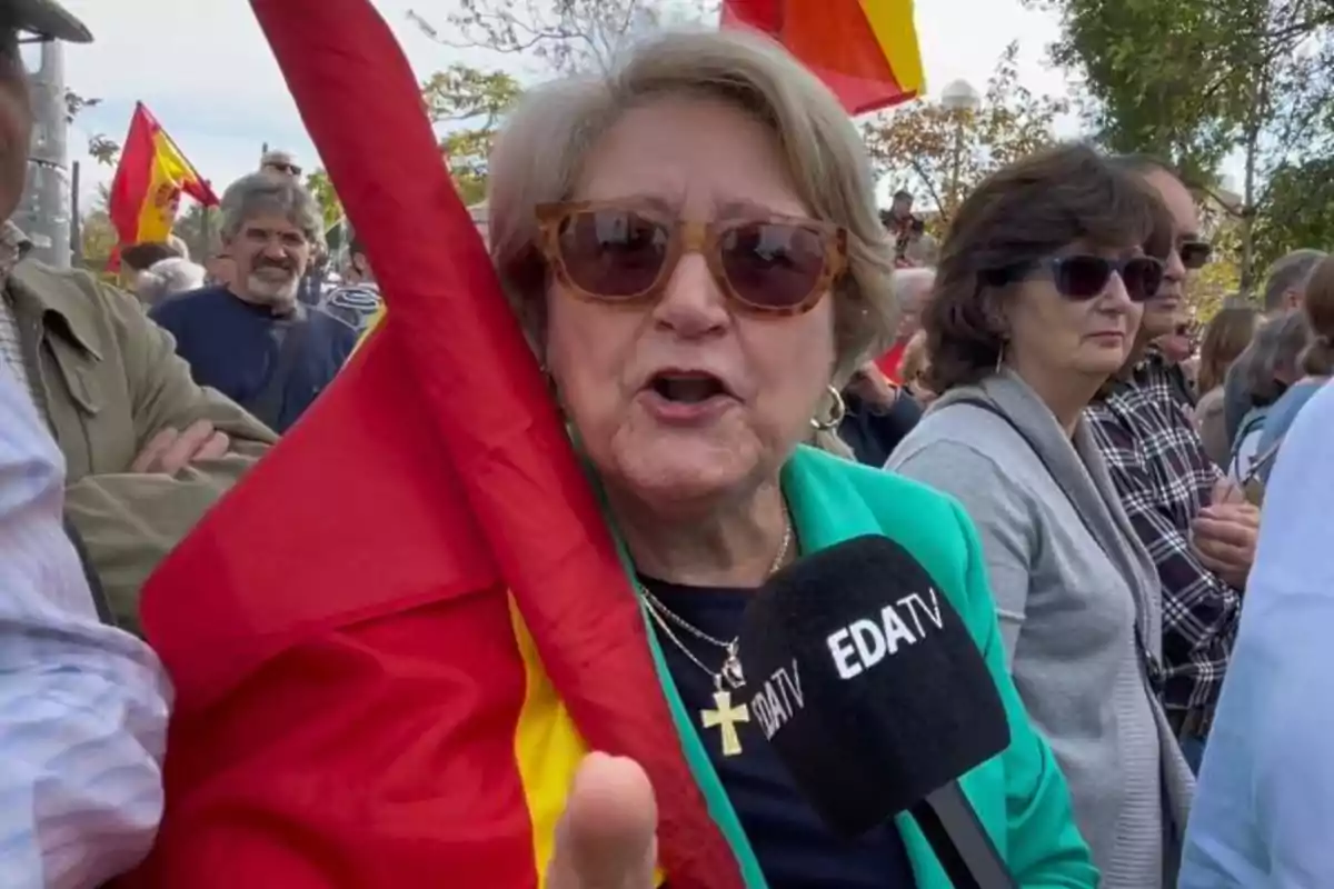 Una mujer con gafas de sol sostiene una bandera mientras es entrevistada por un micrófono de EDATV en una manifestación.