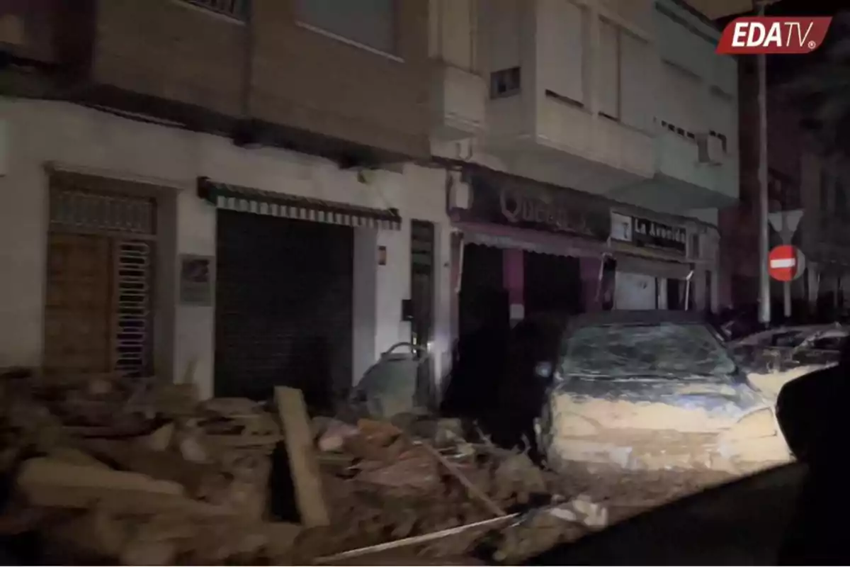 Calle con escombros y un coche cubierto de barro frente a edificios con persianas cerradas durante la noche.