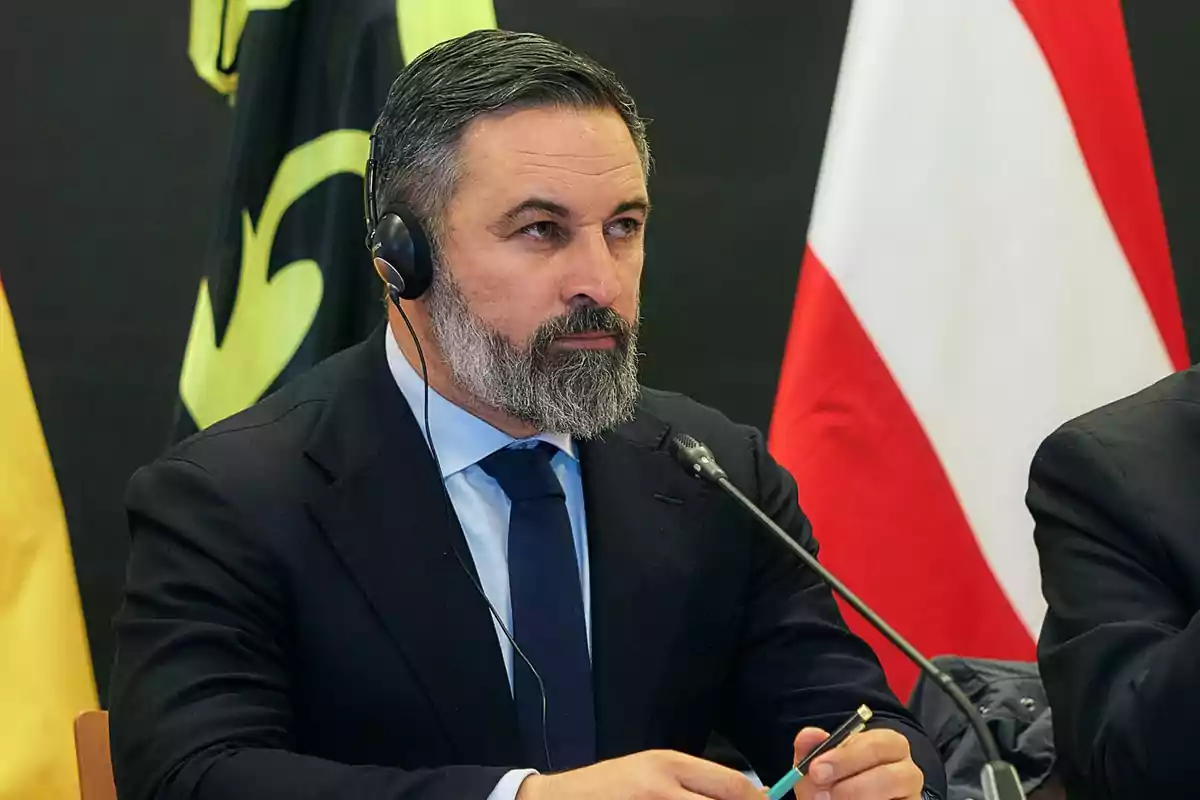 Un hombre con auriculares y barba está sentado en una mesa con micrófono, vistiendo traje y corbata, con banderas de fondo.