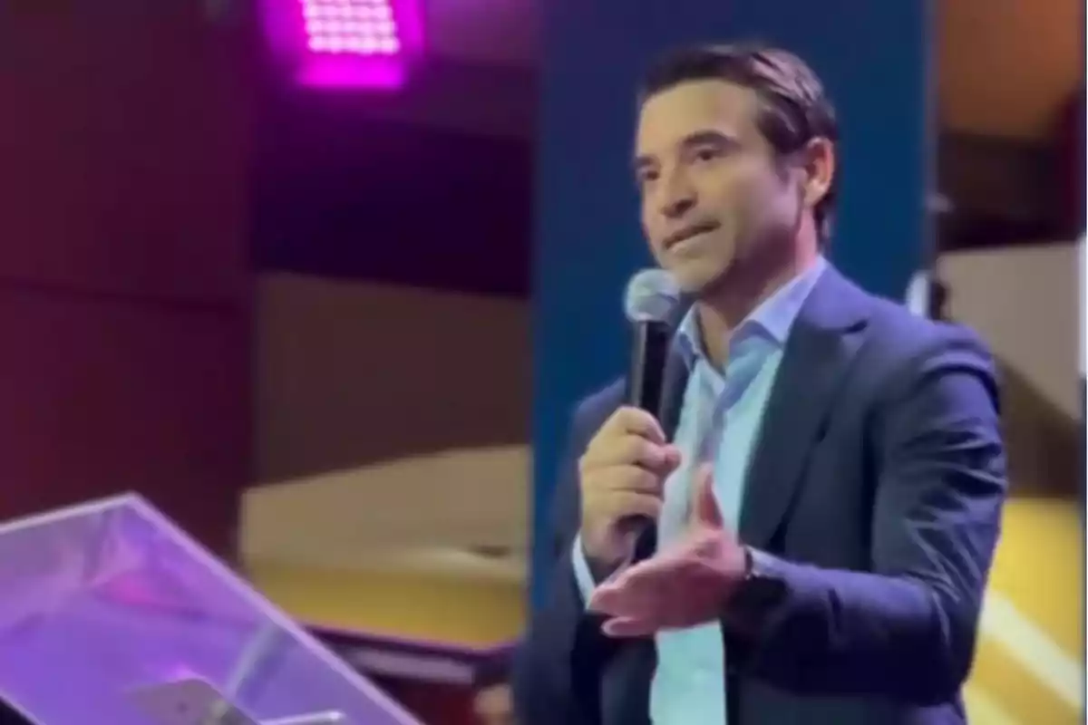 Un hombre en traje hablando en un escenario con un micrófono en la mano.