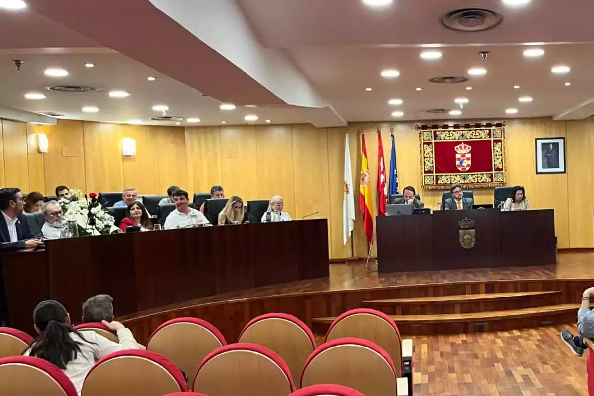 Personas sentadas en un salón de plenos con paredes de madera, banderas y escudo oficial al fondo, algunas personas en el público y otras en la mesa principal Personas sentadas en un salón de plenos con paredes de madera, banderas y escudo oficial al fondo, algunas personas en el público y otras en la mesa principal