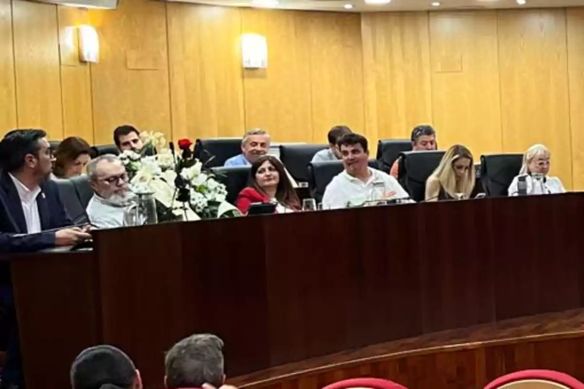 Personas sentadas en una mesa de madera en lo que parece ser una sala de reuniones con paredes de madera y arreglos florales en el centro Personas sentadas en una mesa de madera en lo que parece ser una sala de reuniones con paredes de madera y arreglos florales en el centro
