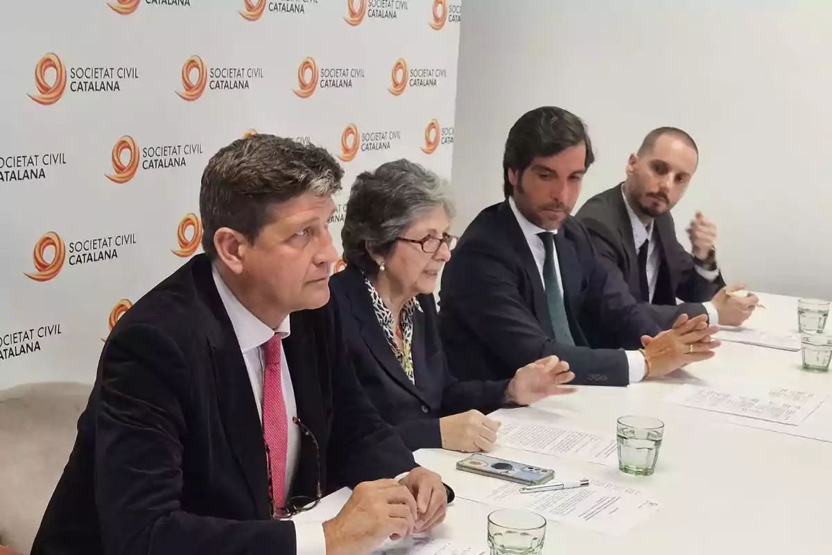Cuatro personas con ropa formal están sentadas en una mesa durante una reunión de trabajo con papeles y vasos de agua, al fondo se ve un panel con el logo de Societat Civil Catalana.