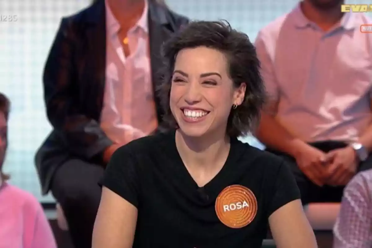 Rosa sonriente con camiseta negra y un gafete naranja que dice Rosa participando en un programa de televisión con público al fondo Rosa sonriente con camiseta negra y un gafete naranja que dice Rosa participando en un programa de televisión con público al fondo