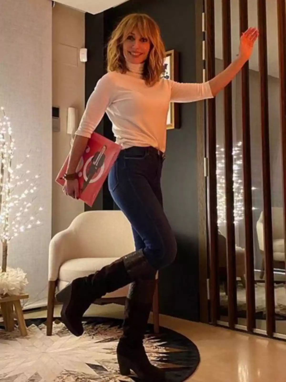 Emma García con cabello rubio y suelto, vestida con un suéter blanco y jeans oscuros, sostiene un libro rojo mientras posa de pie en una habitación decorada con luces y muebles modernos. Emma García con cabello rubio y suelto, vestida con un suéter blanco y jeans oscuros, sostiene un libro rojo mientras posa de pie en una habitación decorada con luces y muebles modernos.