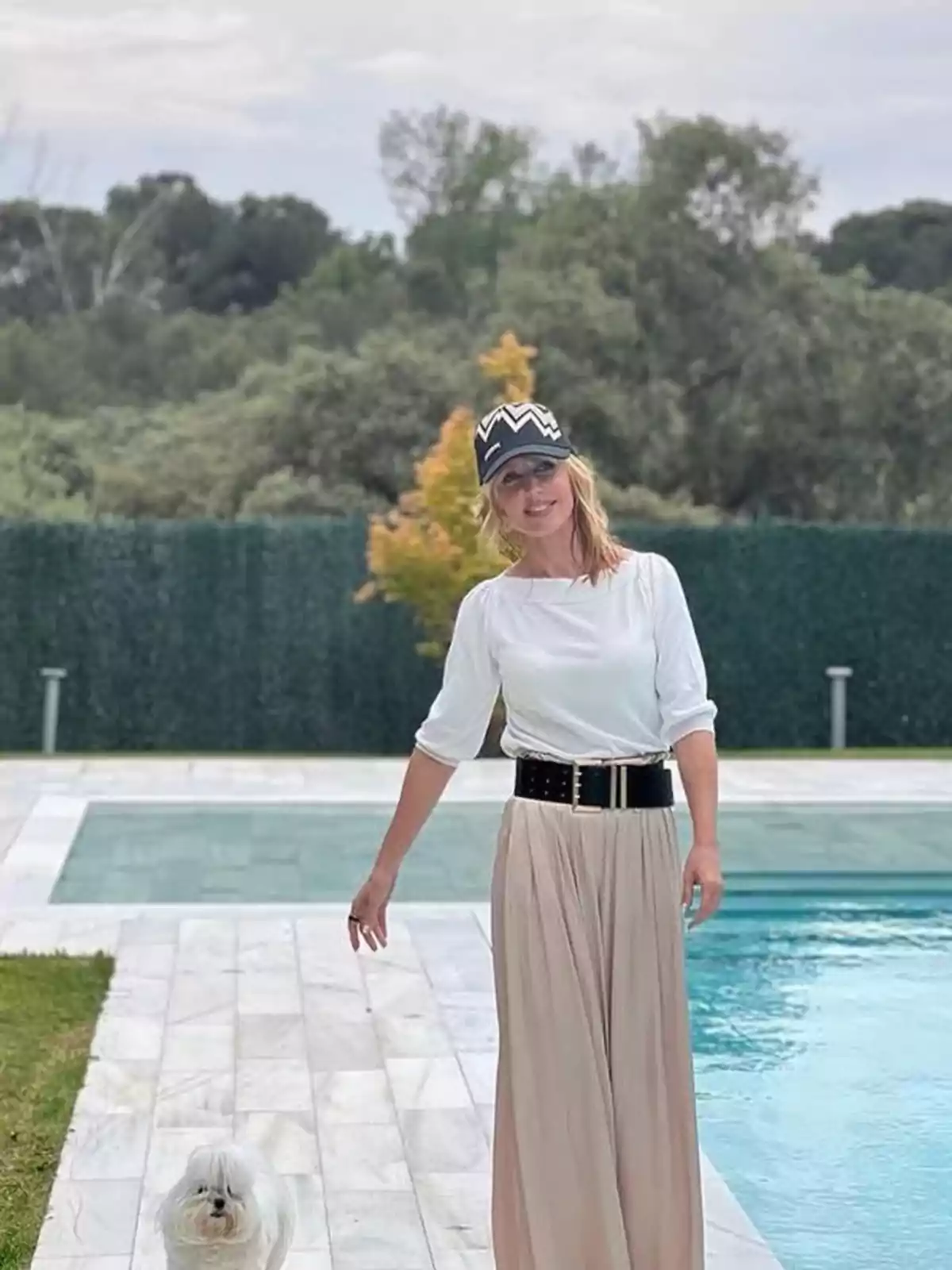 Emma García con sombrero y falda larga está de pie junto a una piscina, acompañada de un perro pequeño, con un fondo de árboles y setos. Emma García con sombrero y falda larga está de pie junto a una piscina, acompañada de un perro pequeño, con un fondo de árboles y setos.