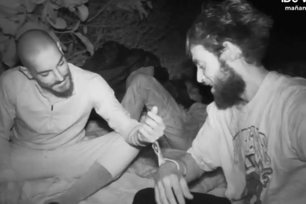 Damián y Montoya sentados en el suelo durante la noche se miran y sonríen mientras uno de ellos sostiene la muñeca del otro Damián y Montoya sentados en el suelo durante la noche se miran y sonríen mientras uno de ellos sostiene la muñeca del otro