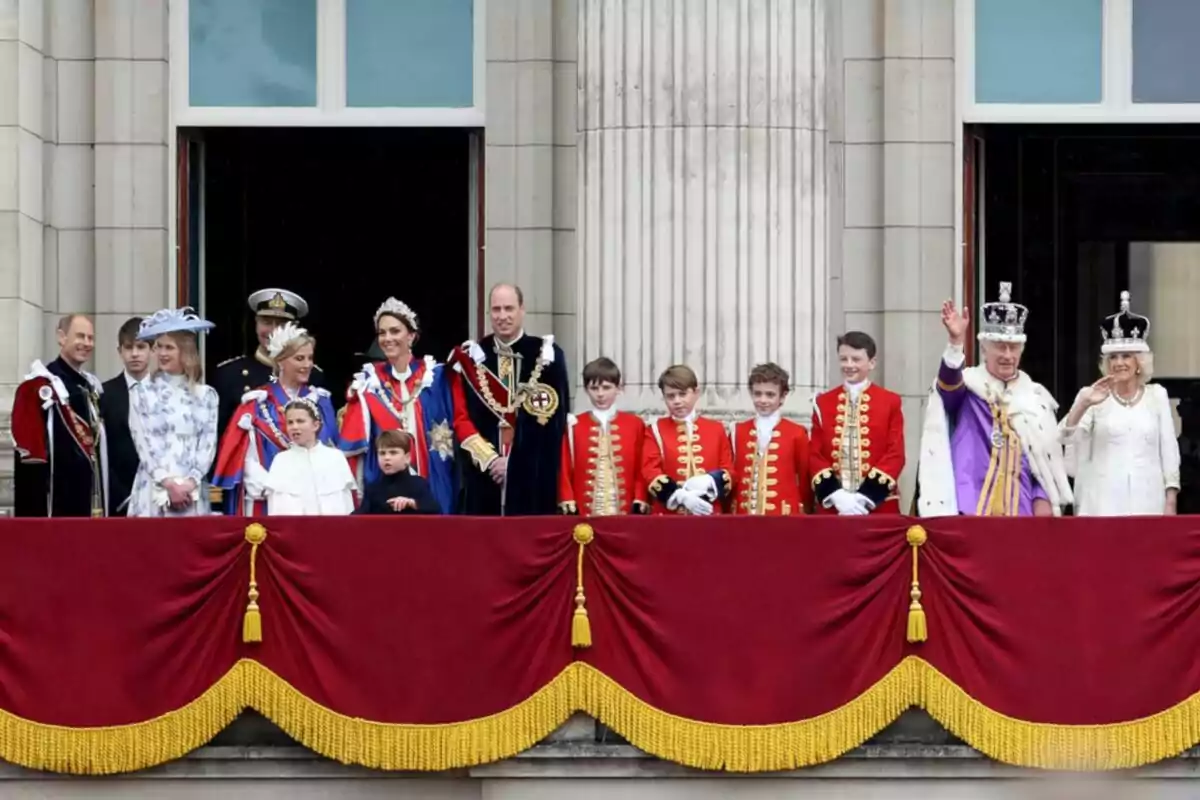 Un grupo de personas vestidas con trajes formales y coloridos se encuentra en un balcón decorado con cortinas rojas y doradas frente a un edificio de columnas. Un grupo de personas vestidas con trajes formales y coloridos se encuentra en un balcón decorado con cortinas rojas y doradas frente a un edificio de columnas.