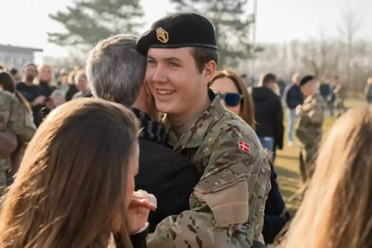 Un joven soldado con uniforme militar sonríe mientras abraza a una persona en un evento al aire libre con más personas alrededor. Un joven soldado con uniforme militar sonríe mientras abraza a una persona en un evento al aire libre con más personas alrededor.