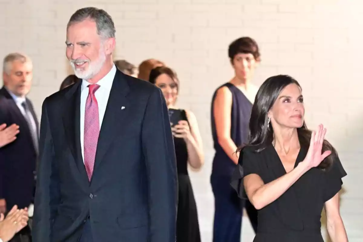 El rey Felipe con traje oscuro y corbata roja sonríe mientras la reina Letizia vestida de negro saluda con la mano, al fondo hay varias personas observando y una de ellas toma una foto con el móvil. El rey Felipe con traje oscuro y corbata roja sonríe mientras la reina Letizia vestida de negro saluda con la mano, al fondo hay varias personas observando y una de ellas toma una foto con el móvil.