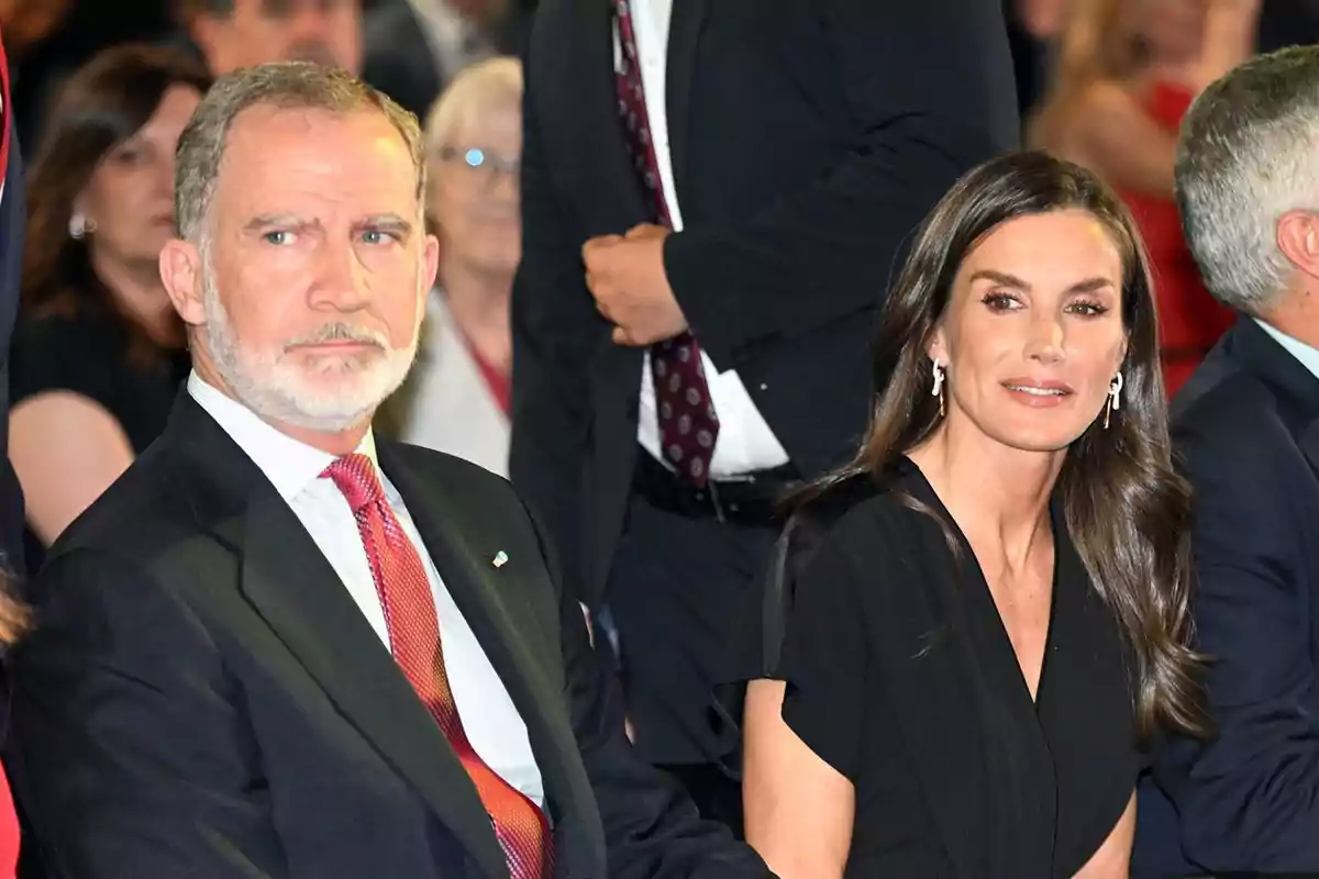 El rey Felipe con barba y traje oscuro junto a la reina Letizia de cabello largo y vestido negro sentados en un evento formal rodeados de otras personas El rey Felipe con barba y traje oscuro junto a la reina Letizia de cabello largo y vestido negro sentados en un evento formal rodeados de otras personas