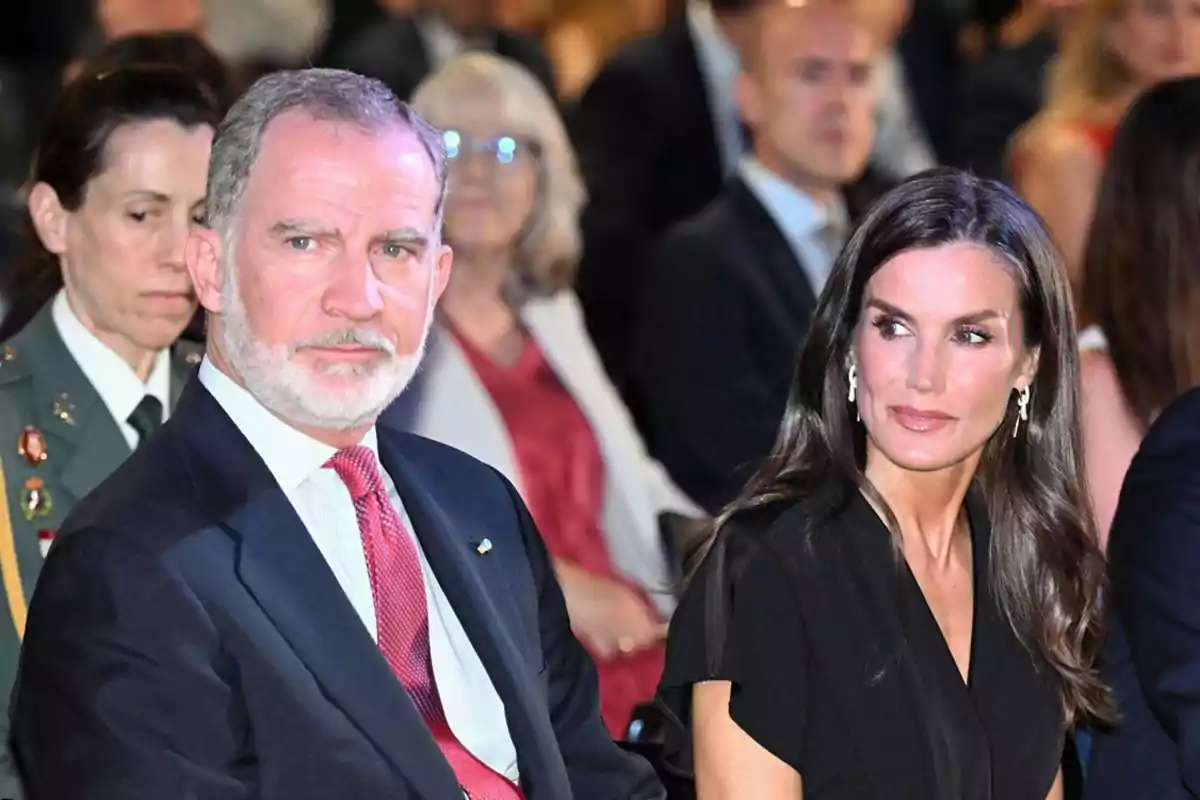 A man in a dark suit and red tie is sitting next to a woman with long dark hair dressed in black at a formal event surrounded by other people. A man in a dark suit and red tie is sitting next to a woman with long dark hair dressed in black at a formal event surrounded by other people.