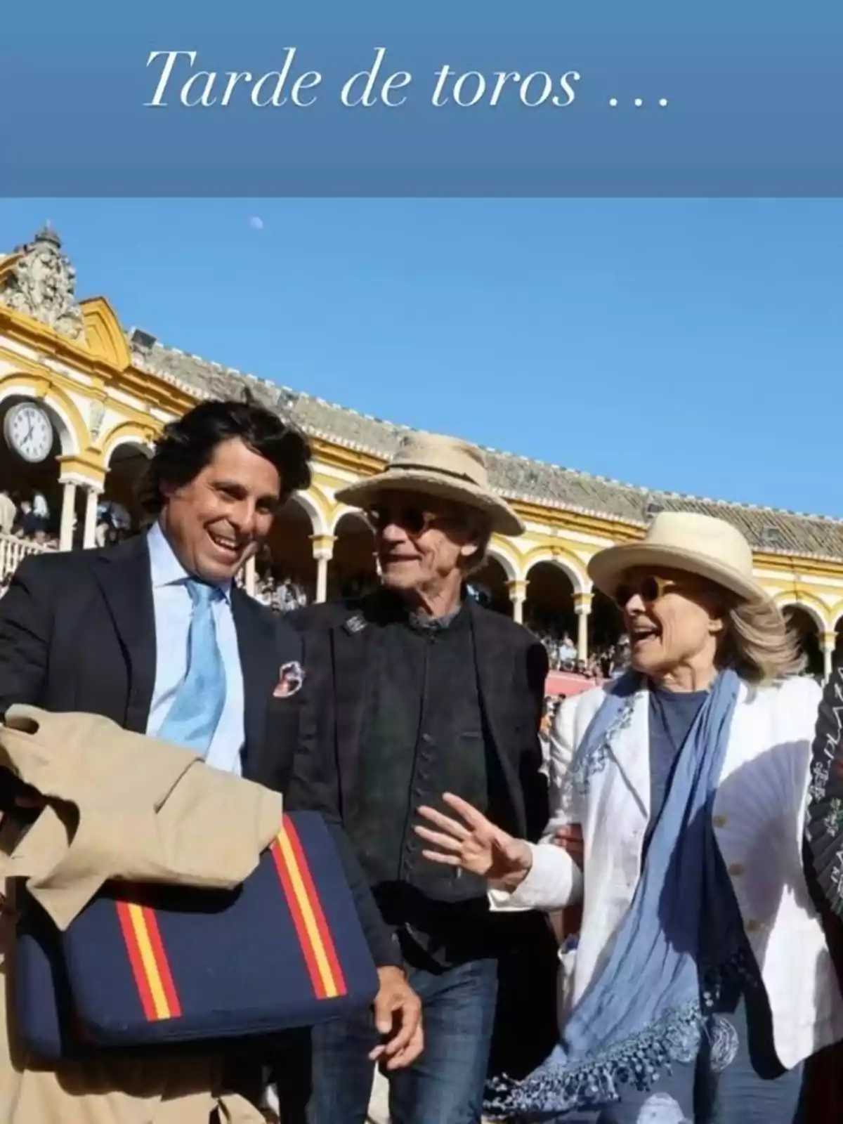 Tres personas sonrientes conversan en una plaza de toros, con un edificio de fondo y un cielo despejado. Tres personas sonrientes conversan en una plaza de toros, con un edificio de fondo y un cielo despejado.