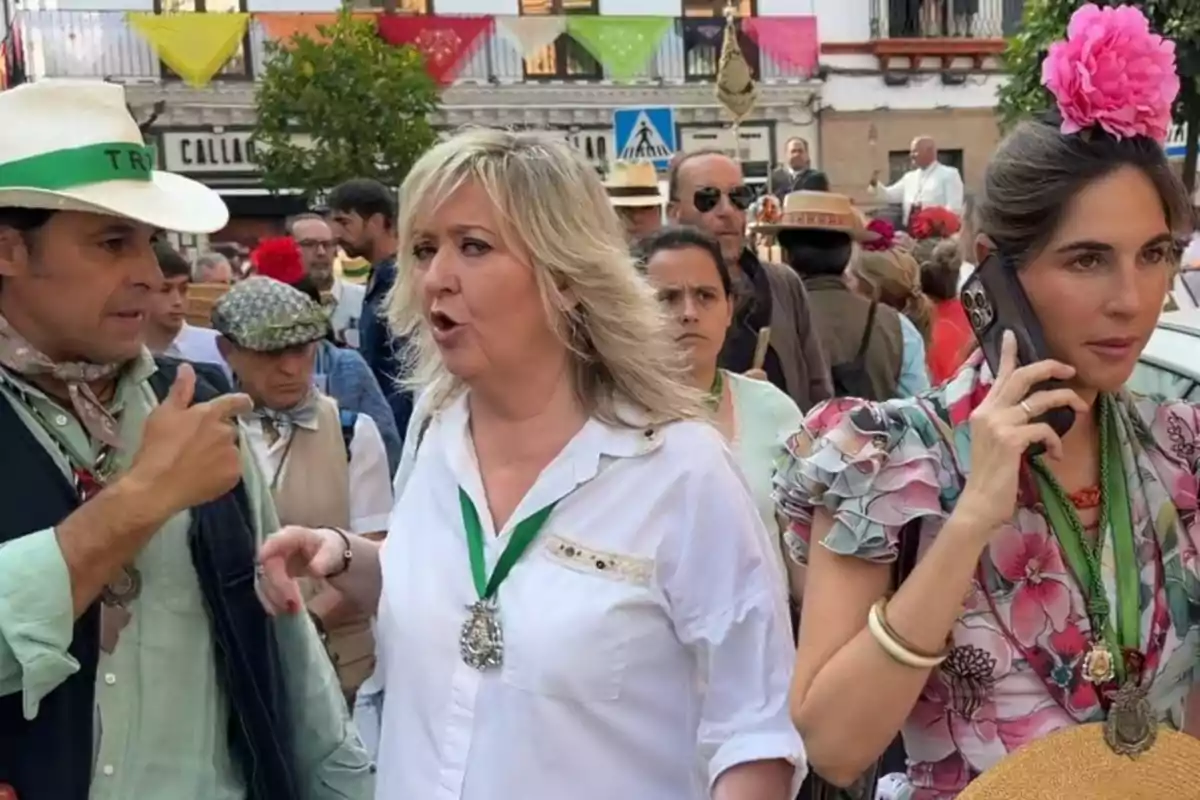Personas vestidas con trajes típicos andaluces participan en una celebración en la calle, una mujer lleva una flor rosa en el cabello y habla por teléfono, mientras otras personas conversan y caminan entre decoraciones coloridas. Personas vestidas con trajes típicos andaluces participan en una celebración en la calle, una mujer lleva una flor rosa en el cabello y habla por teléfono, mientras otras personas conversan y caminan entre decoraciones coloridas.