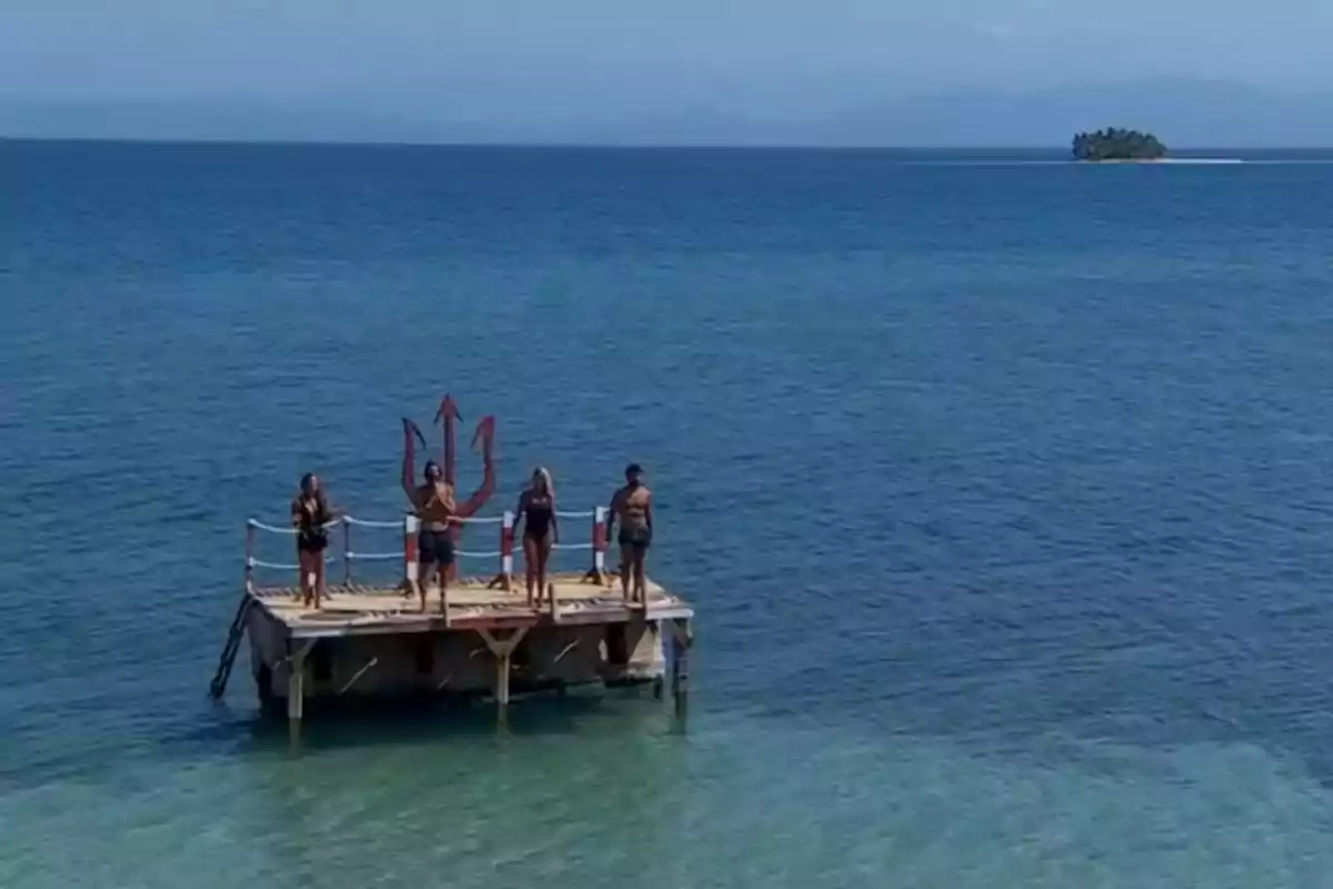 Un grupo de personas está de pie sobre una plataforma en el mar con un tridente grande detrás de ellos y una pequeña isla se ve a lo lejos. Un grupo de personas está de pie sobre una plataforma en el mar con un tridente grande detrás de ellos y una pequeña isla se ve a lo lejos.