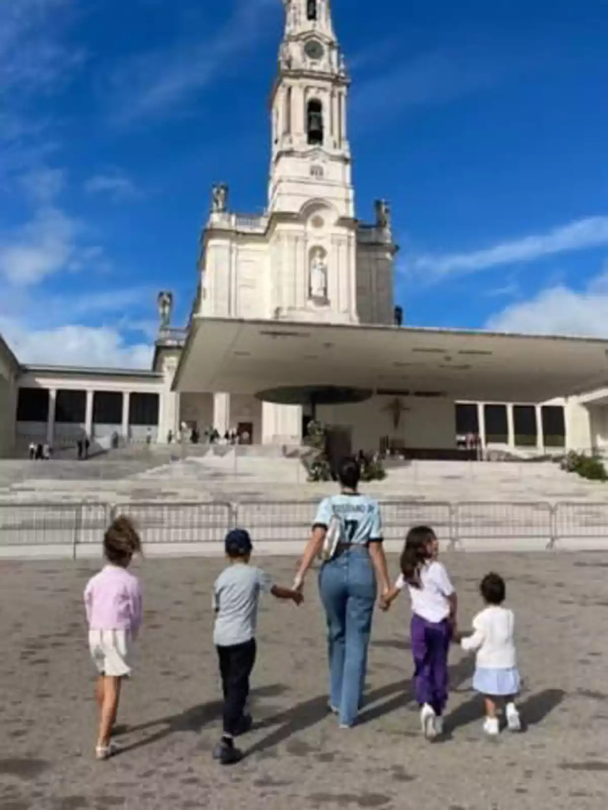 A woman and four children walk hand in hand toward a religious building with a tall tower under a blue sky. A woman and four children walk hand in hand toward a religious building with a tall tower under a blue sky.